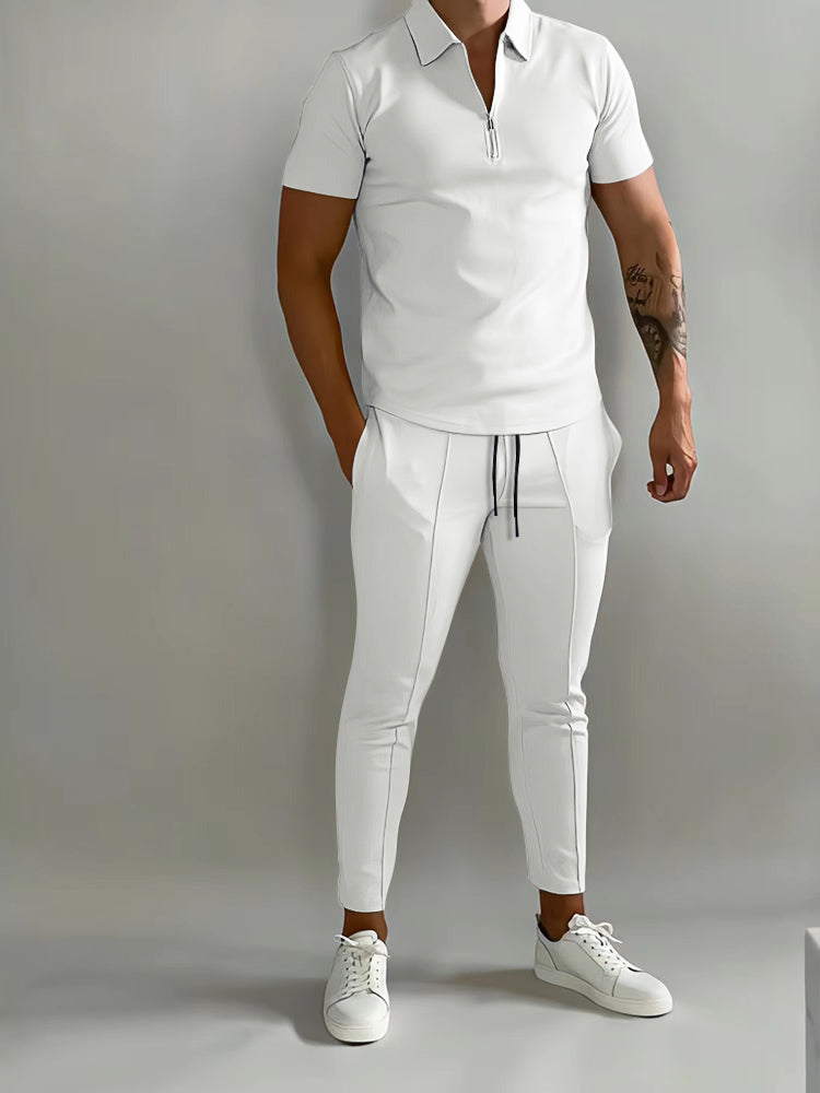 De afbeelding toont een man in een stijlvol wit trainingspak van het merk Opestico. Het pak heeft een moderne pasvorm, met een korte rits op de bovenkant en sportieve details zoals de trekkoord aan de broek. De man combineert het pak met witte sneakers, wat zorgt voor een frisse, elegante uitstraling. Het trainingspak biedt zowel comfort als stijl, ideaal voor een casual look of sportieve activiteit.