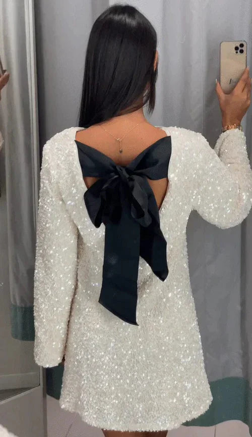 odel draagt de Ophira Dreamwear Elsa Glitter Jurk, een sprankelende witte jurk met een elegante zwarte strik aan de achterkant, terwijl ze zichzelf in de spiegel bewondert.