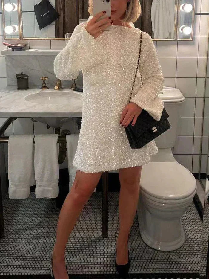 Model in de Ophira Dreamwear Elsa Glitter Jurk in wit, bedekt met glinsterende pailletten, terwijl ze poseert in een badkamer met een Chanel tas en zwarte hakken, met een spiegelreflexie in de achtergrond.