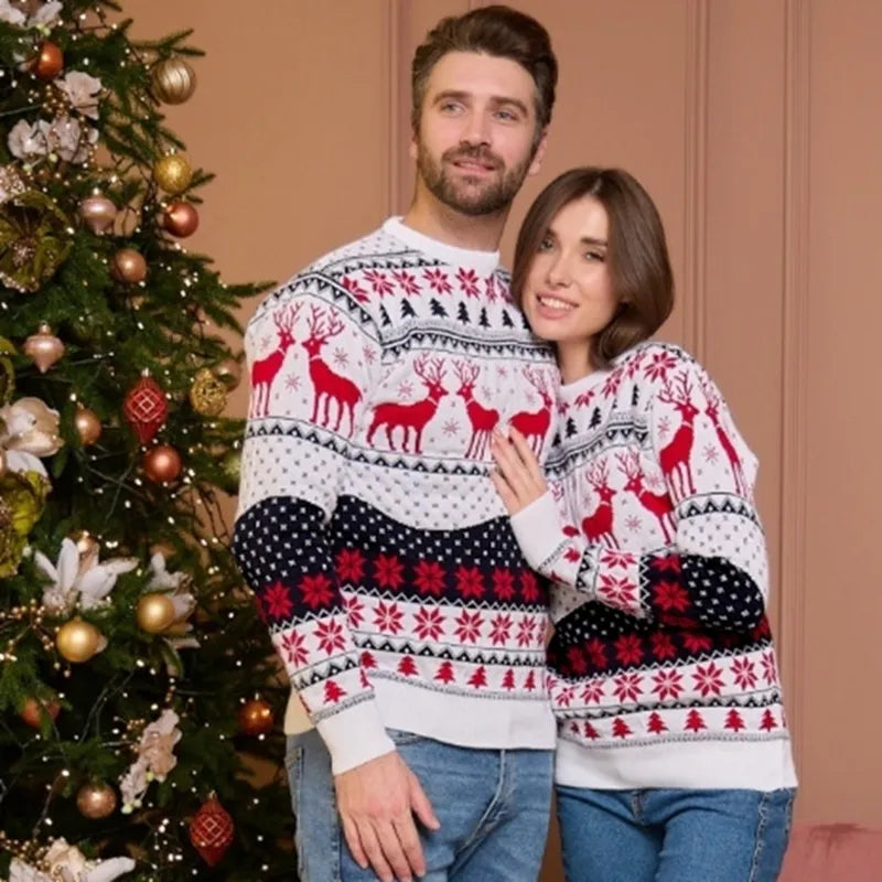 Een man en een vrouw dragen een bijpassende kersttruienset met een feestelijk patroon van rendieren, sneeuwvlokken en kerstachtige motieven in rood, zwart en wit. Ze staan samen voor een kerstboom, wat de gezellige kerststemming benadrukt. De truien zijn zowel stijlvol als comfortabel.