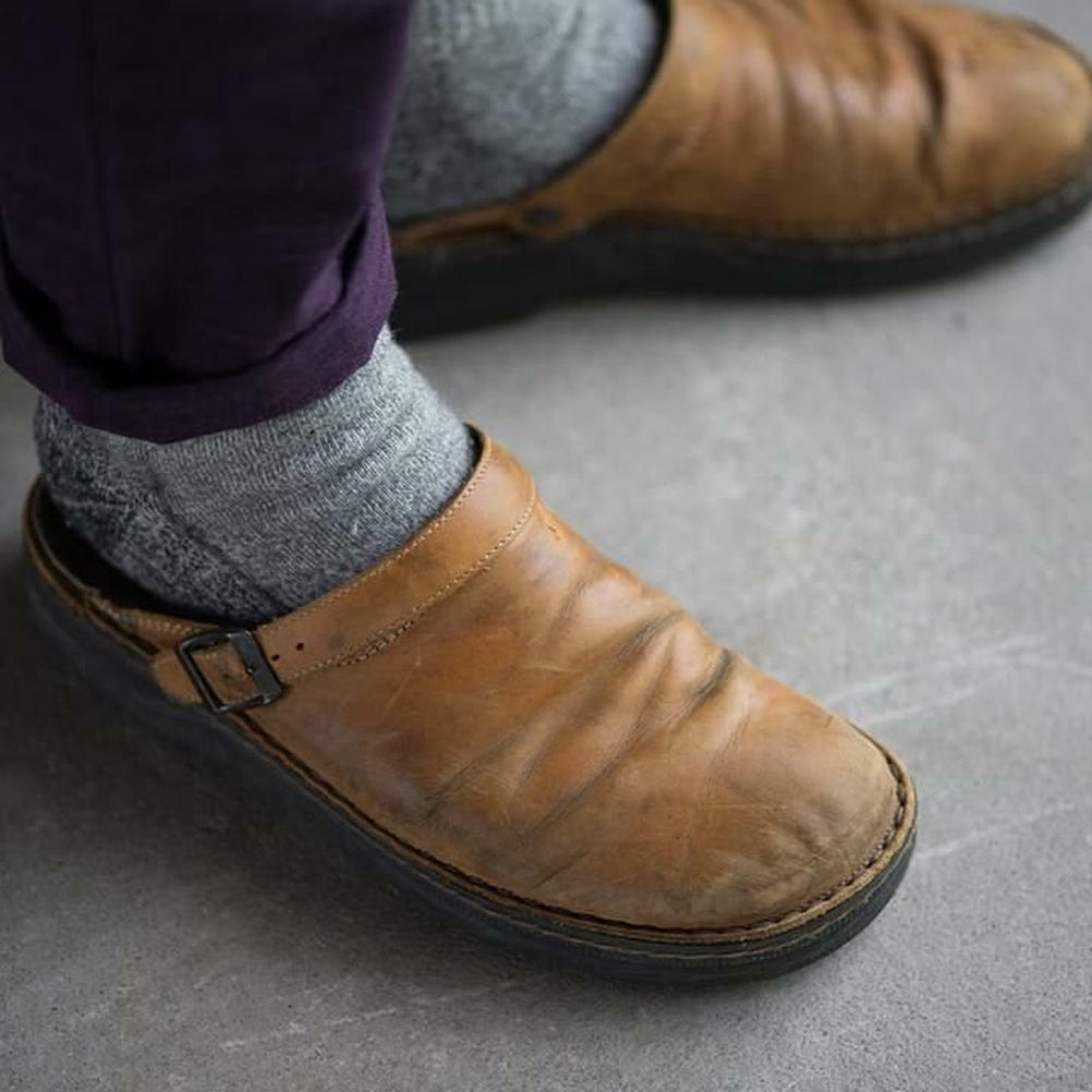 Bruine leren slippers van Paxano, gedragen met grijze sokken en een paarse broek. Deze slippers combineren comfort en stijl, ideaal voor dagelijks gebruik met een verstelbare band voor een perfecte pasvorm.