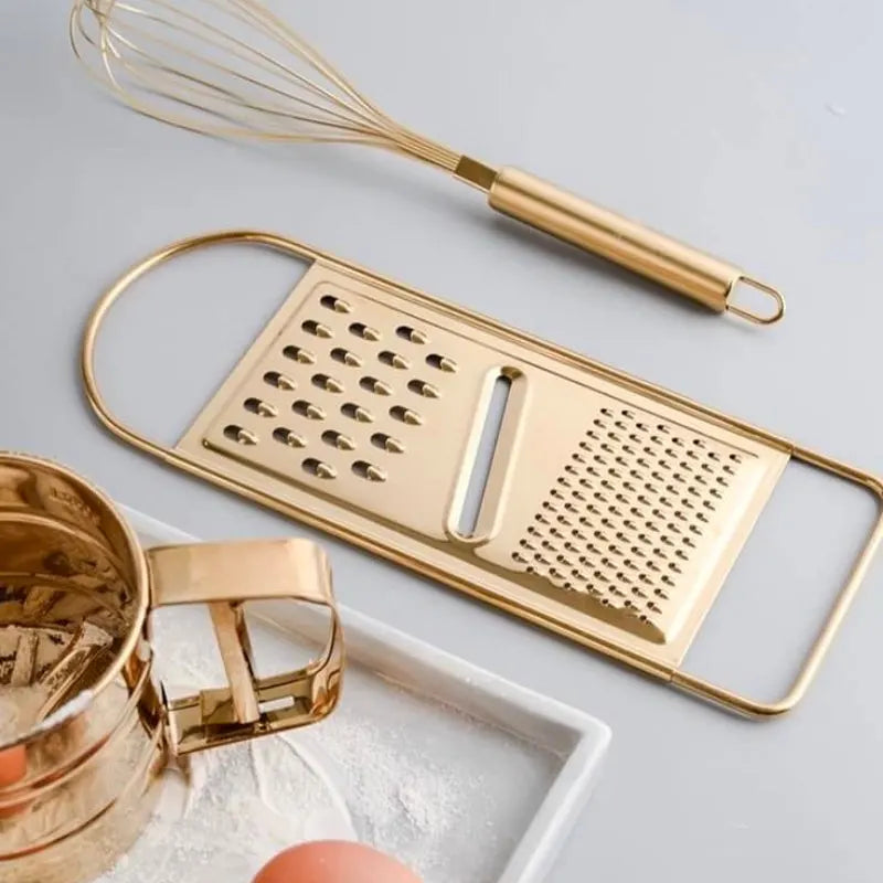 Een gouden vlakrasp, een garde en een zeef liggen op een werkblad naast eieren en bloem. De rasp heeft meerdere scherpe gaatjes voor efficiënt schillen en raspen van ingrediënten. De stijlvolle keukengerei zijn ideaal voor het bereiden van maaltijden en brengen een vleugje elegantie in je keuken. Het is een veelzijdig keukengerei voor dagelijks gebruik.
