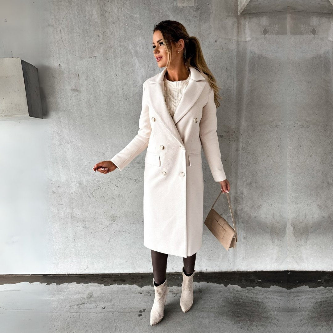 Ulara Winterjas - Een elegante lange dames winterjas in gebroken wit met een stijlvolle pasvorm, dubbele rij knopen en brede revers. Gecombineerd met een chique handtas en enkellaarsjes voor een complete, verfijnde look.