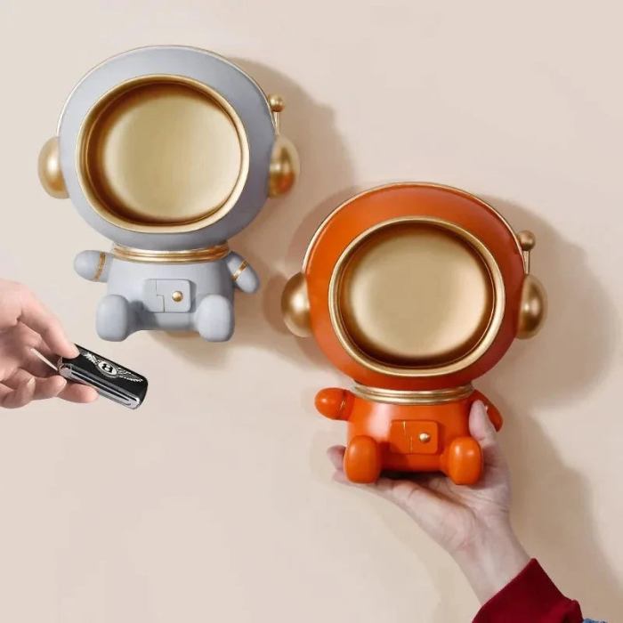 Twee schattige astronaut-sleutelhangers, één in grijs en één in oranje, beide met een gouden helm. De sleutelhangers zijn functioneel en kunnen aan de muur worden bevestigd, ideaal voor het ophangen van sleutels, oordopjes of andere kleine voorwerpen. Het ontwerp is speels en decoratief, perfect voor een moderne en functionele ruimte.