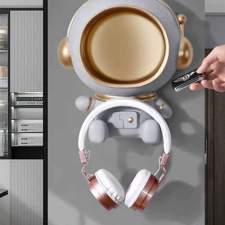 Een schattige sleutelhanger in de vorm van een astronaut met een gouden helm en witte koptelefoon. De sleutelhanger is functioneel en kan aan de muur worden bevestigd, perfect voor het ophangen van sleutels, oordopjes of andere kleine voorwerpen. Het is een decoratief en praktisch item voor elke ruimte, met een modern en speels ontwerp.