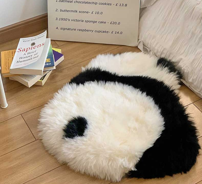 Een zwart-wit panda-vormig vloerkleed op een houten vloer, naast een stapel boeken. Het zachte, harige kleed biedt bescherming aan de vloer en voegt een speelse en comfortabele sfeer toe aan de kamer. Het ontwerp van het kleed met een panda-gezicht maakt het een uniek en charmant accessoire voor de ruimte.
