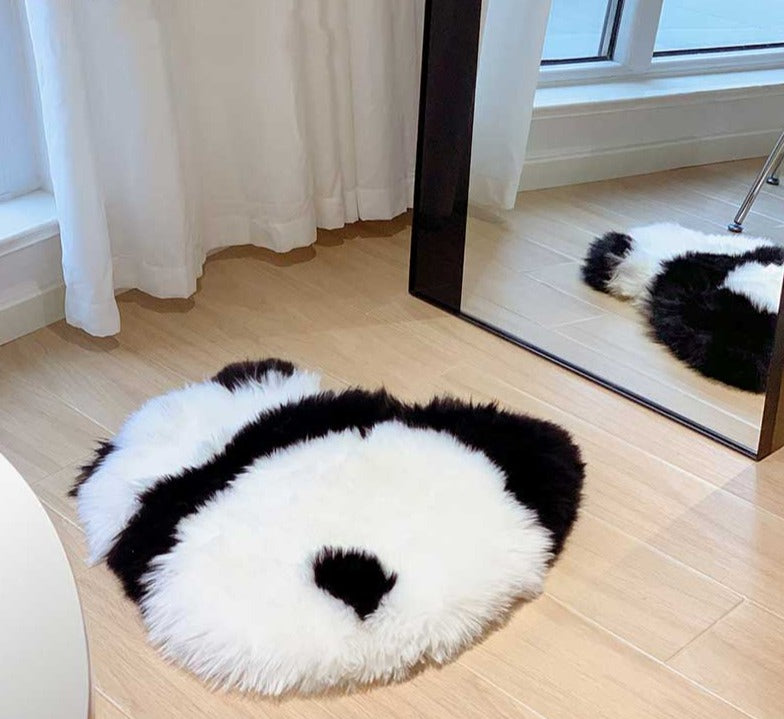 Een schattig zwart-wit panda-vormig vloerkleed in een moderne kamer, perfect voor bescherming van de vloer. Het zachte, harige matje ligt op een houten vloer, vlakbij een spiegel, en biedt zowel comfort als stijl aan de ruimte. Het ontwerp van het kleed voegt een speelse en unieke uitstraling toe aan de kamer.