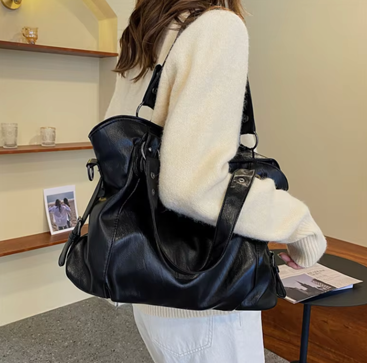 Zyrano zwarte leren crossbody tas, praktisch en stijlvol, gedragen over de schouder. De tas heeft een ruime en flexibele vorm, ideaal voor dagelijks gebruik, en is voorzien van stevige handvatten en een schouderriem. De vrouw draagt de tas casual met een beige trui, wat de veelzijdigheid van het ontwerp benadrukt. Perfect voor elke gelegenheid, van werk tot weekend.